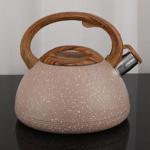 Puede incluir: Una tetera moteada de color topo con asa y tapa de madera. El asa es arqueada y la tapa tiene un pomo de madera a juego. La tetera tiene un pico plateado.