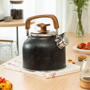 Könnte beinhalten: Schwarz gesprenkelte Emaille-Teekanne mit Holzgriff und Deckelknopf. Die Kanne hat einen Ausguss aus Edelstahl und steht auf einem weiß-rot gestreiften Tuch. Ein klassischer Küchenartikel.