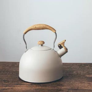 May include: A cream-colored stovetop kettle with a wooden handle and spout detail. The kettle has a rounded body and a silver base. The handle and spout accents are made of light-colored wood. The kettle sits on a wooden surface.