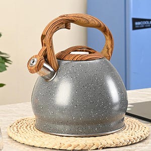 May include: A gray speckled tea kettle with a wooden handle and lid. The kettle has a silver spout and sits on a woven straw mat. The handle is arched and made of light brown wood.