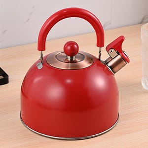 May include: A red stovetop kettle with a curved red handle and a red knob on the lid. The kettle has a copper-colored base and spout. The kettle is sitting on a wooden surface.