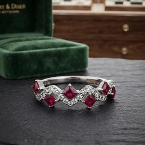 May include: A silver-toned ring featuring a repeating pattern of princess-cut red rubies and small round white stones. The band has a zigzag or chevron-inspired design with alternating gemstone settings, creating a continuous metallic structure.