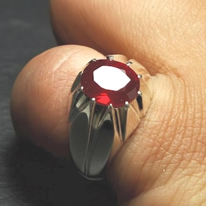 Peut inclure: Une bague en argent ornée d'un grand rubis ovale et facetté. Le rubis est d'un rouge profond et vibrant. Le sertissage de la bague présente un motif gravé détaillé sur les côtés.