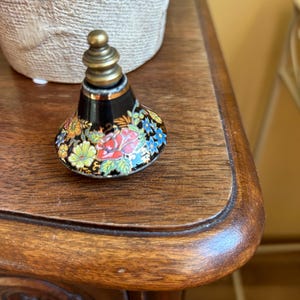 Op de afbeelding: Een zwarte flesstopper met een bloemmotief en een messing bovenkant. De stopper heeft een kleurrijk patroon van rode, blauwe, gele en groene bloemen. Hij ligt op een houten oppervlak.
