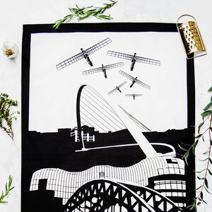 May include: Black and white tea towel featuring a graphic illustration of the Newcastle upon Tyne skyline. The design includes the Tyne Bridge, the Sage Gateshead, and the Angel of the North sculptures. The words "BALTIC FLOUR MILLS" are printed on the towel.