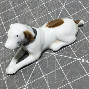 Puede incluir: Figura de cerámica de un perro blanco con manchas marrones y un collar negro. El perro está tumbado sobre una superficie gris con líneas de cuadrícula. Las orejas y la espalda del perro están manchadas de marrón.