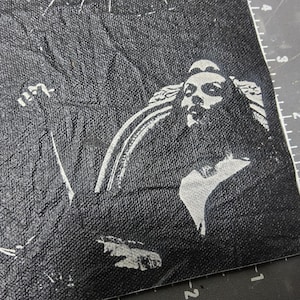 May include: A black fabric patch featuring a high-contrast white screen-printed graphic of a figure with long hair and a detailed collar. The material has a textured, slightly wrinkled surface, and a portion of a ruler is visible along the edge.