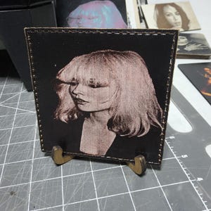 May include: A square, black leather coaster with a sepia-toned portrait of a woman with blonde hair. The coaster is displayed on a small wooden stand. Other coasters with similar images are visible in the background.