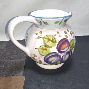 May include: A white ceramic pitcher with a rounded body and a curved handle. The pitcher is decorated with hand-painted purple plums, green leaves, and brown vines. The rim is trimmed with a blue band.