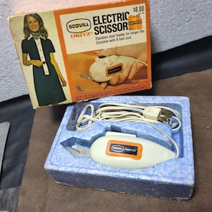 May include: Vintage Scovill Dritz electric scissors in original box. The white and orange scissors are in a blue foam insert. The box features an illustration of the scissors and a woman in a dark dress. The cord is 8 feet long.
