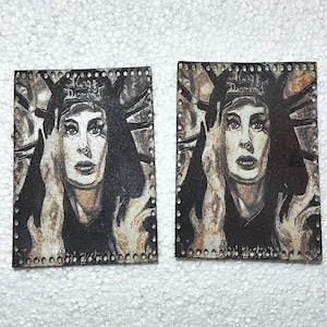 May include: Two rectangular leather patches featuring a portrait of a woman with long hair and a crown. The patches have a distressed, vintage look with the text "King Diamond" at the top. The edges are perforated with small holes.