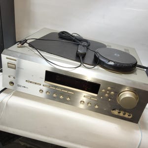 Pode incluir: Receptor de áudio prateado com um leitor de CD preto e uma antena preta. O receptor tem vários botões e um visor digital. Um cabo preto conecta o leitor de CD ao receptor.