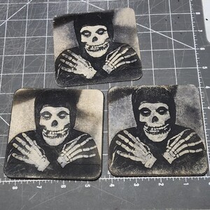 May include: Three square coasters featuring a black and white image of a skull-faced figure with skeleton hands. The figure is wearing a black hooded outfit. The coasters are arranged on a gray surface.