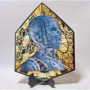 May include: A blue and black decorative art piece featuring a portrait of a character with pins in their head. The artwork is set within a geometric frame with yellow and black patterns. The piece is displayed on a black stand.