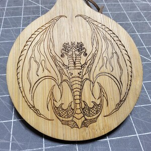 May include: A round, wooden ornament with a dragon design etched into the surface. The dragon has large wings and a detailed body. A leather cord is attached for hanging. The ornament is made of light-colored wood.