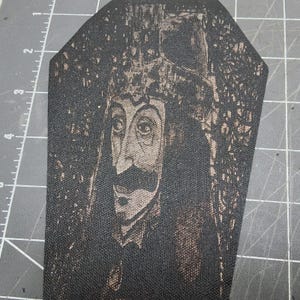May include: A coffin-shaped fabric patch featuring a black and copper-toned screen-printed portrait of a man with a mustache and a tall hat. The patch is positioned on a cutting mat with visible inch markings along the left side for scale.