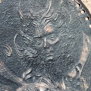 May include: A circular black metal plaque featuring a raised relief design of a face with stylized hair or horns. The surface has a rough, textured finish with hints of copper-colored oxidation visible in the recessed areas and along the edges.