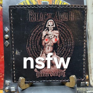 May include: Black leather patch with a graphic of a skeletal figure in a nun's habit, with red star-shaped pasties. The text "BLACK ARIA II" and "glenn danzig" are visible. The word "nsfw" is overlaid on the image.