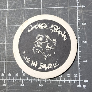 May include: A round, black and white coaster with the text "Concrete Danks Live in Brazil" in a stylized font. The center features a skeleton graphic. The coaster has a white border and is likely made of absorbent material.