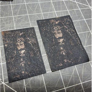 Può includere: Due toppe rettangolari in tessuto nero con un ritratto color seppia di un uomo con baffi e abiti ornati. Le toppe sono tagliate da un pezzo di tessuto più grande e poste su un tappetino da taglio grigio.