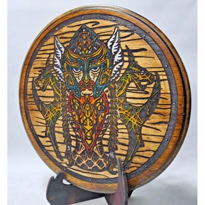 Puede incluir: Arte mural redondo de madera con un diseño detallado de guerrero vikingo. La obra de arte presenta un casco colorido, barba y motivos de dragones, con un borde marrón oscuro y un fondo de vetas de madera natural. La pieza se exhibe sobre un soporte de madera.