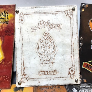 May include: White leather wallet featuring the band Motörhead's logo and the phrase "Ace of Spades." The design includes a skull within a diamond shape. The wallet is rectangular with metal studs in each corner.