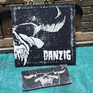 May include: Two black patches with white graphics. The larger patch features a skull with horns and the word "DANZIG" in white. The smaller patch has a similar skull design. Both patches have a distressed, vintage look.