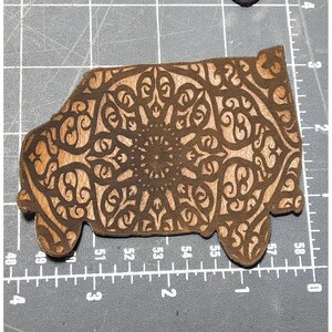 May include: A wooden van-shaped ornament with an intricate mandala design. The van is brown with a lighter wood interior. The ornament is approximately 2.5 inches long and 1.5 inches tall.