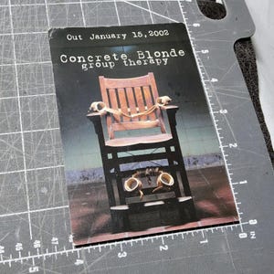 May include: A promotional card for Concrete Blonde's album "group therapy." The card features a wooden chair with restraints and the text "Out January 15, 2002." The chair is the central focus, with a dark background.