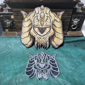 Könnte beinhalten: Zwei Holzausschnitte mit einem detaillierten, stilisierten Monsterdesign. Der größere Ausschnitt ist hellbraun mit dunkelbraunen Akzenten, während der kleinere schwarz mit weißen Details ist. Sie liegen auf einer grünen Oberfläche.