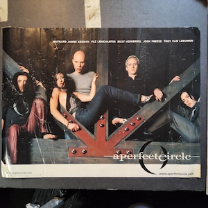 May include: A Perfect Circle band poster featuring the band members posing on a wooden structure with a red arrow. The band's name is displayed in a stylized font. The poster includes the band members' names and website.
