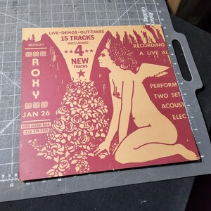 Puede incluir: Una portada de álbum de estilo vintage con un esquema de color rojo y beige. El diseño presenta una figura desnuda con alas de ángel, un gran ramo de rosas y el texto "ROXY" y "15 Tracks". La portada del álbum también incluye la fecha "JAN 26".