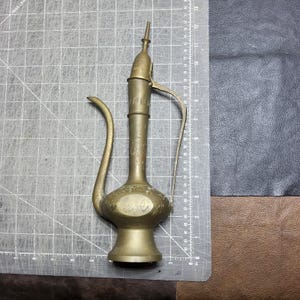 Pode incluir: Uma jarra de latão metálico com um pescoço alongado, um bico curvo e uma pega fina. A superfície está decorada com padrões florais intrincados e volutas gravadas. Mede aproximadamente 30 cm de altura sobre uma base circular.
