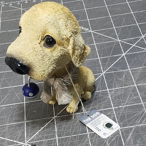 May include: A light tan dog figurine with a black nose and large, dark eyes. The dog is holding a small blue ball with white dots. A tag is attached to the figurine. The dog is sitting on a gray surface.