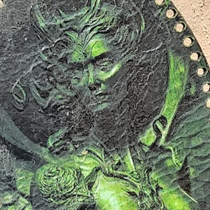May include: A circular wall plaque featuring a textured, relief-style portrait of a figure with flowing hair. The surface is finished in a dark charcoal black with vibrant green highlights accentuating the facial features and hair. The edge has holes.