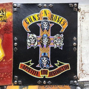 May include: Black leather-bound album cover featuring the Guns N' Roses logo and the words "Appetite for Destruction." The cover has a cross design with skull illustrations. Silver studs are placed around the edges.