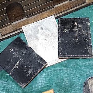 May include: Three small, rectangular, leather-like items with images of skeletons. One is silver, and two are black. The images appear to be vintage or antique. The items are arranged on a green surface.
