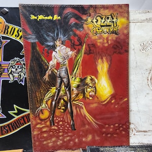 May include: Leather-bound album cover featuring the Ozzy Osbourne album "The Ultimate Sin." The artwork depicts a woman with long black hair and a demon-like figure against a red and yellow background. The Ozzy Osbourne logo is visible.