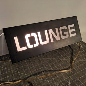 May include: Black rectangular sign with the word "LOUNGE" illuminated in white. The sign has a simple, modern design, with the letters cut out to allow light to shine through. A power cord extends from the sign.