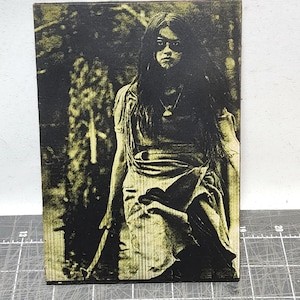 Puede incluir: Placa de madera con una imagen en tonos sepia de una persona con cabello largo y oscuro, vestida con una prenda drapeada y sosteniendo un bastón. El fondo sugiere un entorno forestal. La imagen tiene una estética vintage y artística.