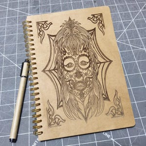 May include: A spiral-bound notebook with a tan cover featuring a laser-engraved sugar skull design of a person with intricate facial patterns and a web-like background, accompanied by a matching tan pen with a black cap and clip.