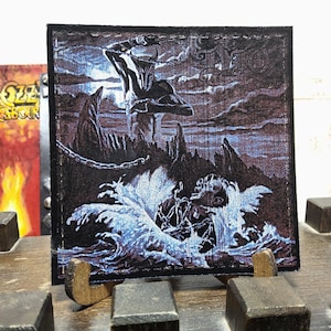 May include: Square patch with a dark, dramatic illustration. A figure in armor raises a weapon over a person in water. The scene is rendered in shades of blue, gray, and black, with a chain and ominous clouds.