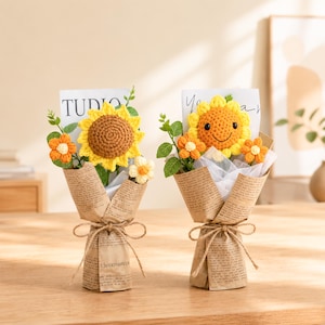 May include: Two crochet sunflower bouquets. Each bouquet features a large yellow sunflower with a brown centre, surrounded by smaller orange and white flowers and green leaves. The bouquets are wrapped in newspaper-style paper and tied with twine.
