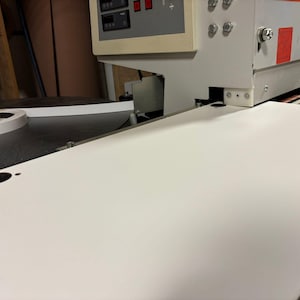 Puede incluir: Primer plano de una pieza rectangular de madera blanca en una máquina para trabajar la madera. La máquina tiene un panel de control con botones y pantallas. Se muestra el proceso de canteado, con un rollo de canto blanco.