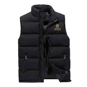 Puede incluir: Chaleco acolchado negro con cuello alto y cremallera completa. Cuenta con dos bolsillos con cremallera y un logotipo dorado que dice "URBAN TRAEDZ". El diseño sin mangas ofrece calidez y estilo.