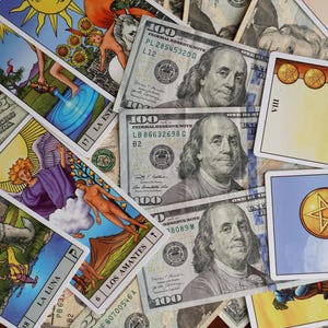 Puede incluir: Una colecci&oacute;n de cartas del tarot con ilustraciones coloridas, incluyendo El Sol, Los Amantes y La Luna, se exhiben junto a varios billetes de cien d&oacute;lares. Las cartas presentan varias figuras y s&iacute;mbolos.