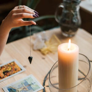 Puede incluir: Una vela blanca encendida en un soporte de vidrio se encuentra sobre una mesa de madera con cartas del tarot. Una mano sostiene un p&eacute;ndulo sobre las cartas. La mesa tiene un dise&ntilde;o de pentagrama.