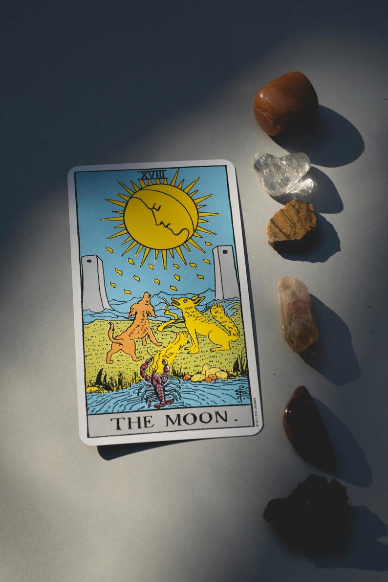 Puede incluir: Una carta del tarot con un sol amarillo con una cara, dos perros y una langosta, con el texto "THE MOON". Varias piedras preciosas pulidas en tonos marr&oacute;n, transparente y naranja est&aacute;n dispersas alrededor de la carta.