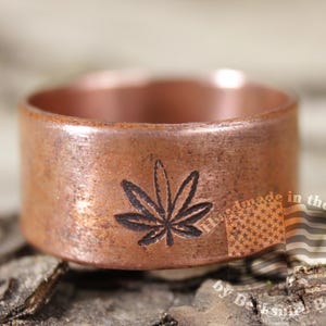 May include: A copper ring with a cannabis leaf design. The ring has a wide band and a rustic finish. The cannabis leaf is etched into the copper. The ring is handmade in the USA.