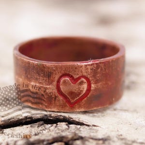 May include: A copper ring with a carved heart design. The ring has a rustic, worn appearance with a reddish-brown hue. The words "Handmade in the USA" are visible on the side of the ring.